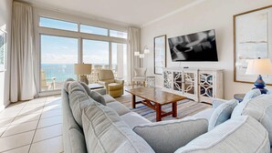 Ein geräumiges Wohnzimmer mit einer großen Couch, einem Couchtisch und einem Fernseher. Ein Balkon bietet Blick auf den Ozean.