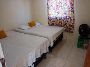Ein Zimmer mit zwei Einzelbetten, jeweils mit weißer Bettdecke und Kissen. Am Fenster befindet sich ein bunter Blumenvorhang und auf dem Boden ein schwarzer Ventilator.