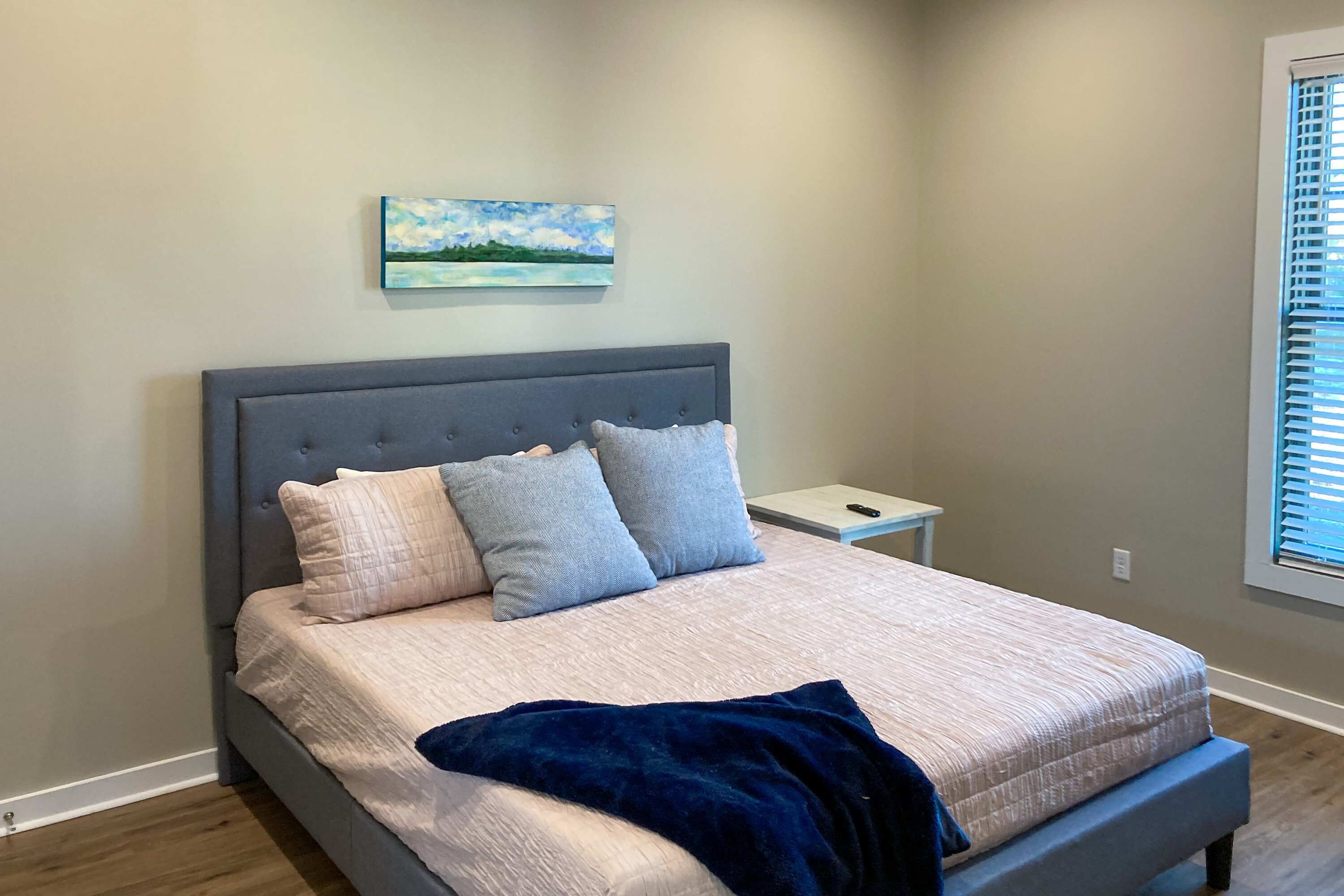 A bedroom with a bed, a nightstand, a painting, and a window with blinds.