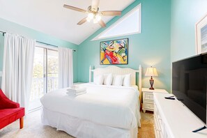 Una camera da letto con un letto, una sedia rossa, una TV, una ventola a soffitto e un quadro appeso al muro.