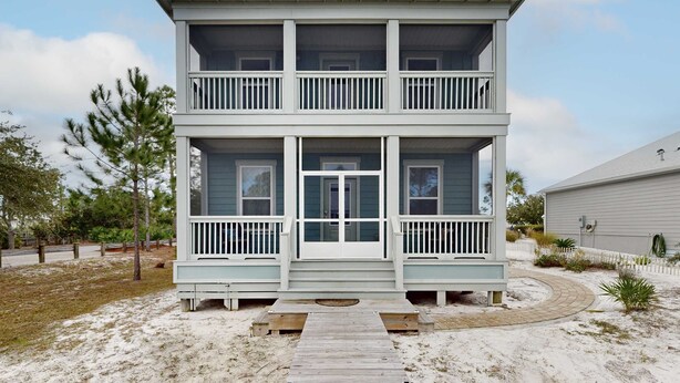Een twee verdiepingen tellend strandhuis met een veranda, een houten terras en een zandpad.