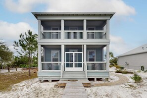 Une maison à deux étages située en bord de mer, dotée d’un porche à l’avant, d’une terrasse en bois et d’une allée menant à la maison, recouverte de sable.