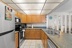 A modern kitchen with wooden cabinets, stainless steel appliances, a granite countertop, and a visible doormat. - Beachfront 2BR Condo w/ Pool & Balcony Views (Panama City Beach)