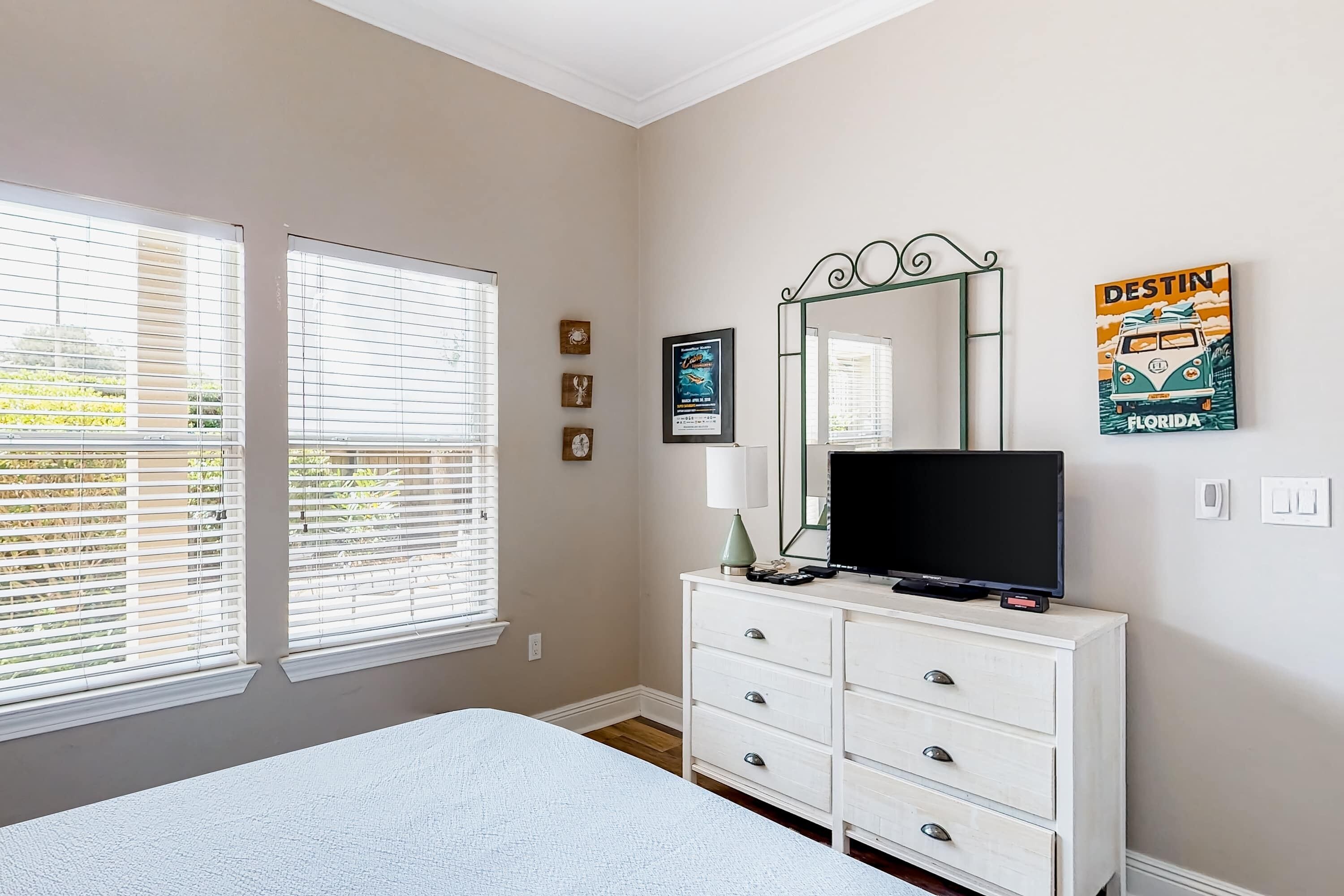 A bedroom with a bed, a dresser, a TV, and a mirror. There are two windows with blinds, a poster on the wall, and a clock.