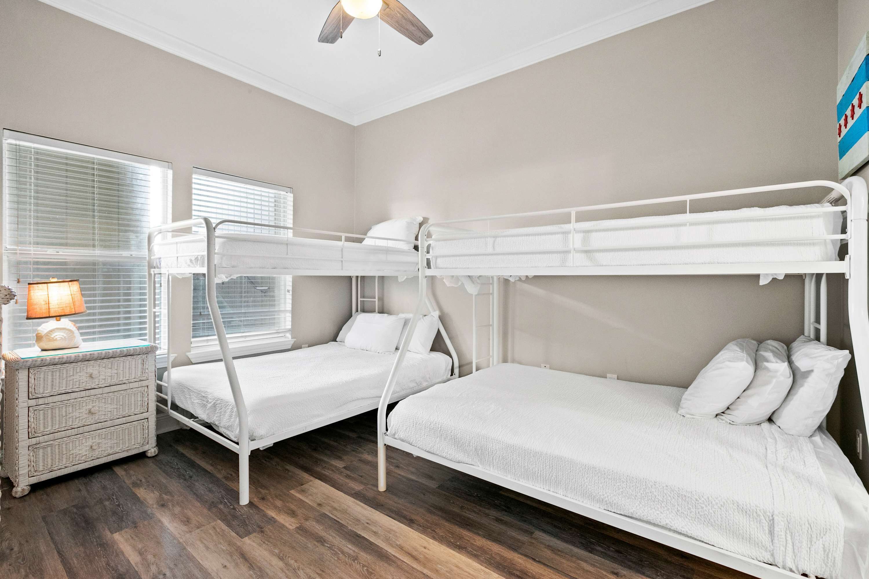 A bunk bed room with a nightstand and lamp, wooden flooring, and a ceiling fan.
