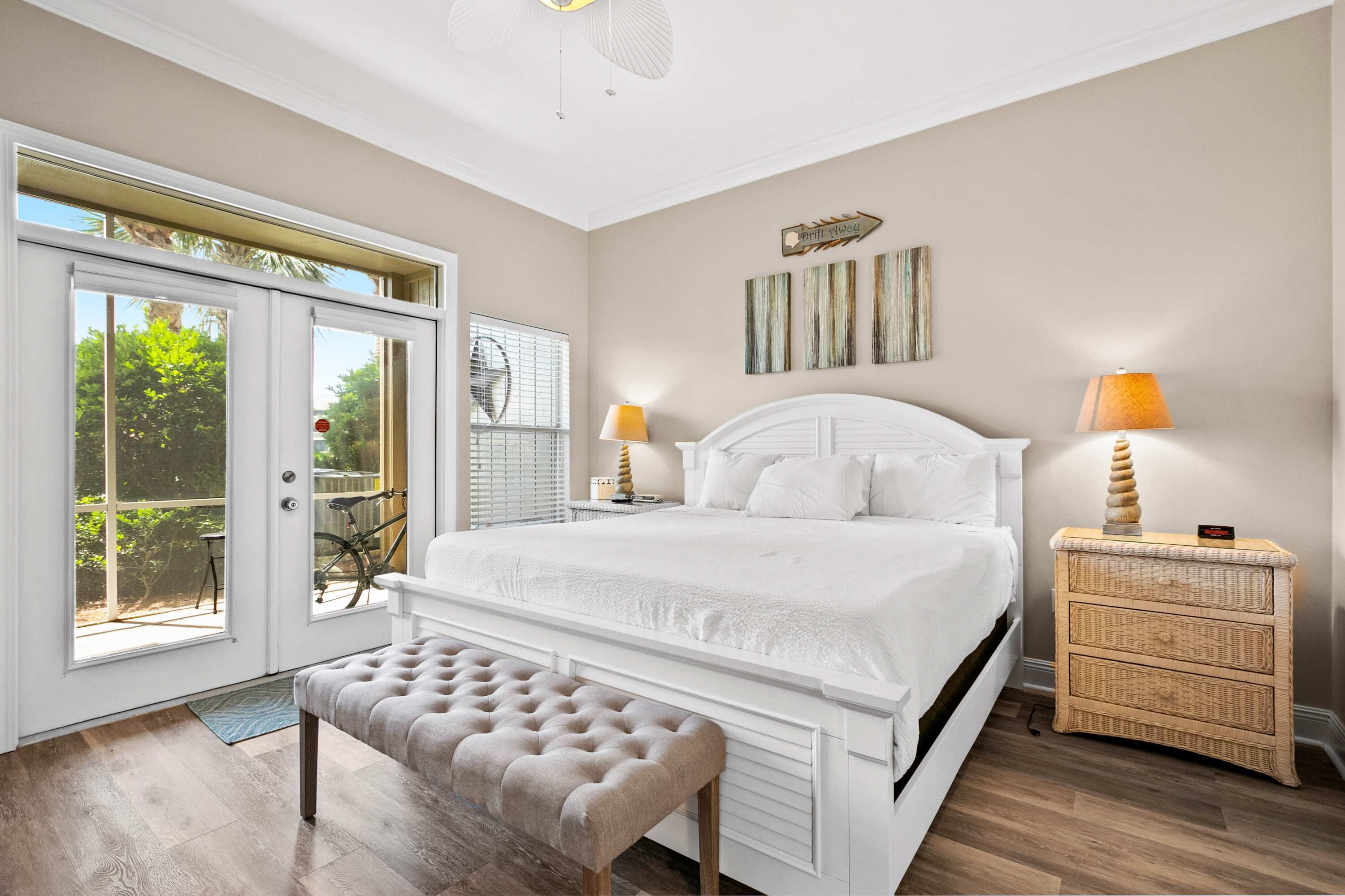 A bedroom with a large bed, a bench, a wicker nightstand, and a view through a sliding glass door.