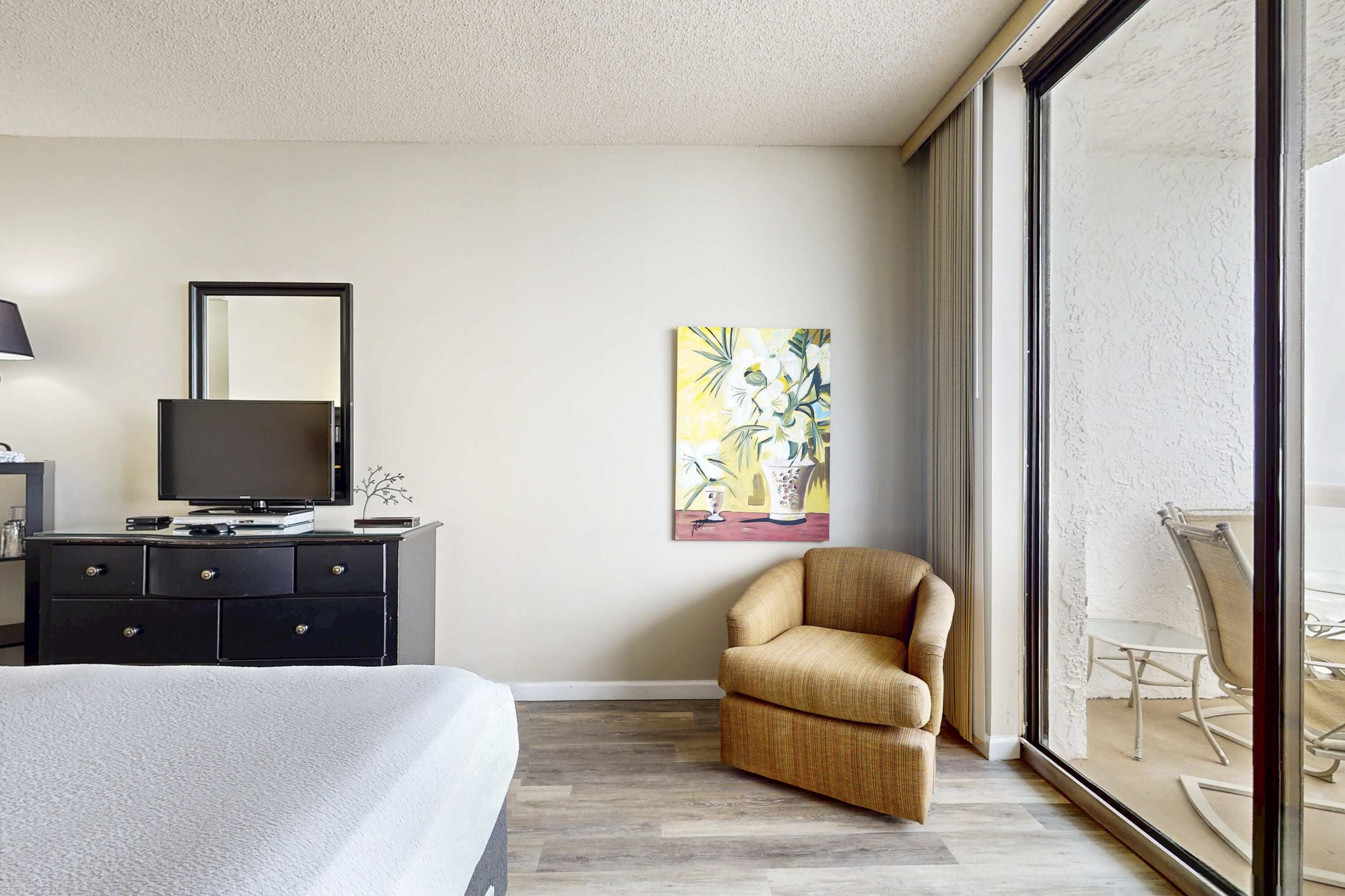 Una camera d'albergo con un letto, una televisione su un comò, una sedia, un quadro appeso al muro e un balcone con tavolo e sedie.