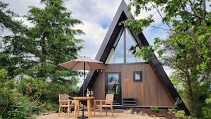 Una cabaña de madera moderna, triangular y con puerta de cristal, en un paisaje natural con árboles y un sendero de grava.