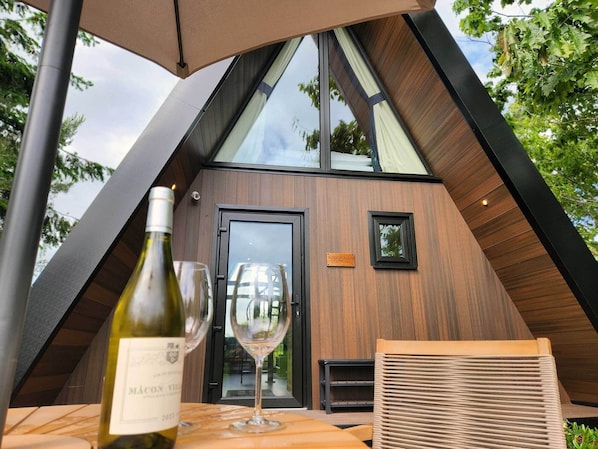 A modern, triangular-roofed building with a wooden exterior and a glass door. A wine bottle and glass are on a table, and a small sign reads "ROCKWALKER LODGE".