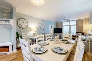 A dining area with a white table set for four, surrounded by white chairs. A large wall clock and framed artwork are visible. - Open & airy 11th-floor condo - close to dining (Destin)
