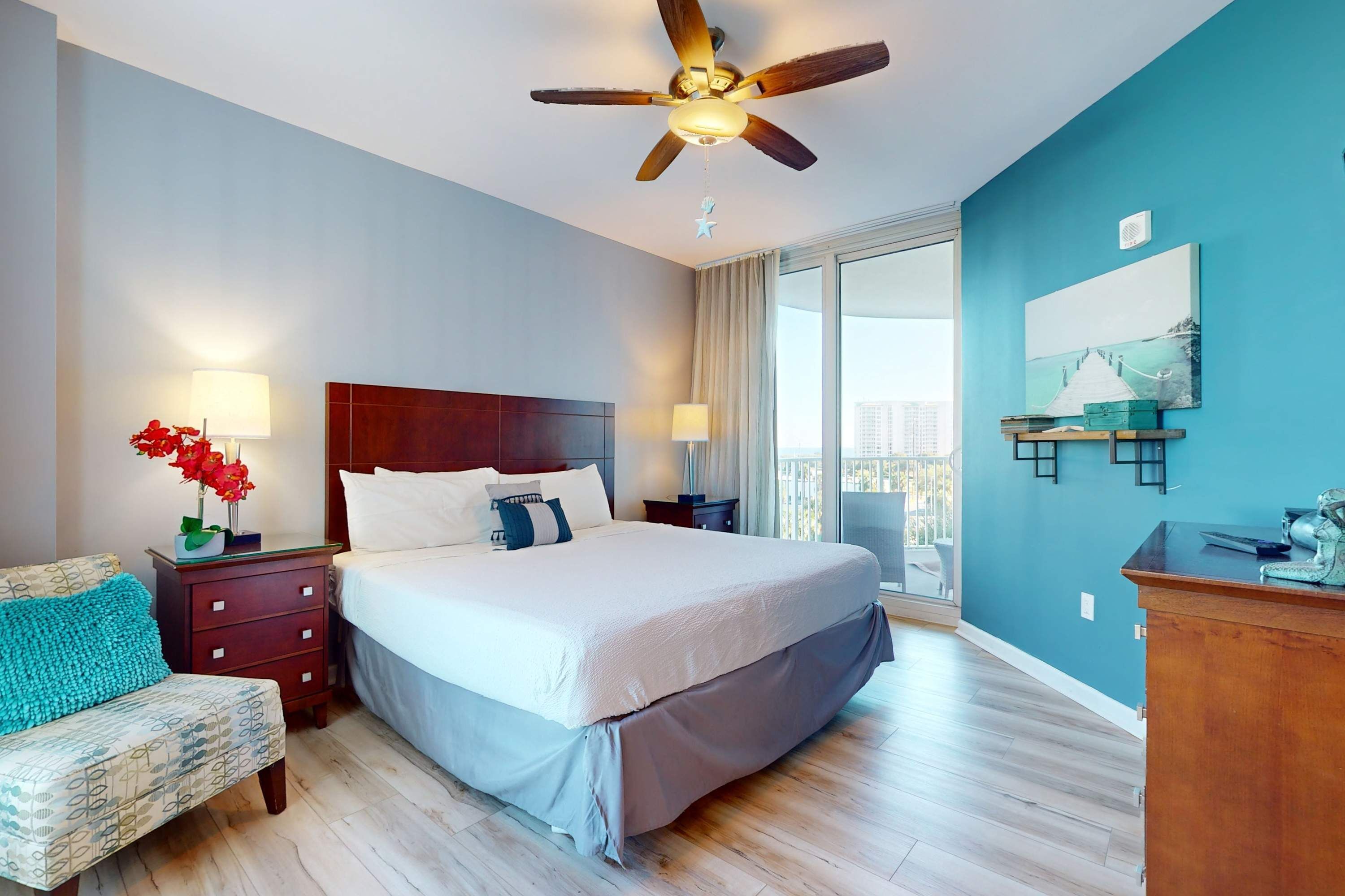 A bedroom with a bed, nightstands, a chair, a ceiling fan, and a view of the outdoors.