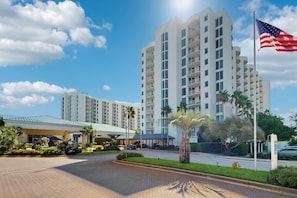 A multi-story hotel with a modern design, featuring balconies on each floor, a landscaped entrance with palm trees, and an American flag. - Coastal condo, shared pools, Onsite eateries, Spa, Wifi (Destin)