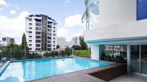 Een modern buitenzwembad met een houten terras, omgeven door een glazen constructie en weelderige begroeiing.