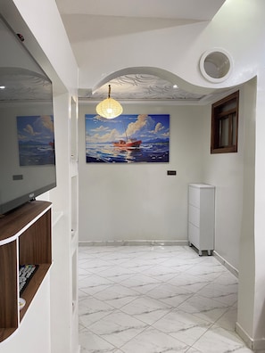 A room with a large painting of a boat on water, a ceiling light, a TV on a wooden stand, and a white cabinet. - 

Amazigh Surf House- Cozy, Quiet, Wi-Fi, Equipped Kitchen . (Aourir, Marruecos)