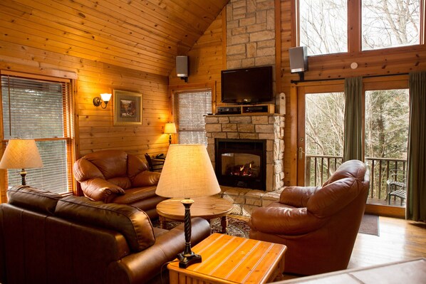Un acogedor interior de cabaña con chimenea, sofás de cuero, una televisión y un balcón con vistas a árboles.
