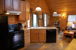 Ein Holzblockhaus-Interieur mit einer Küche, die einen Herd, einen Backofen, eine Spüle und einen Geschirrspüler enthält. Es gibt eine Essecke mit Tisch und Stühlen sowie ein Bücherregal mit Büchern.