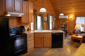 A wooden cabin interior with a kitchen featuring a stove, oven, sink, and dishwasher. There is a dining area with a table and chairs, and a bookshelf with books.
