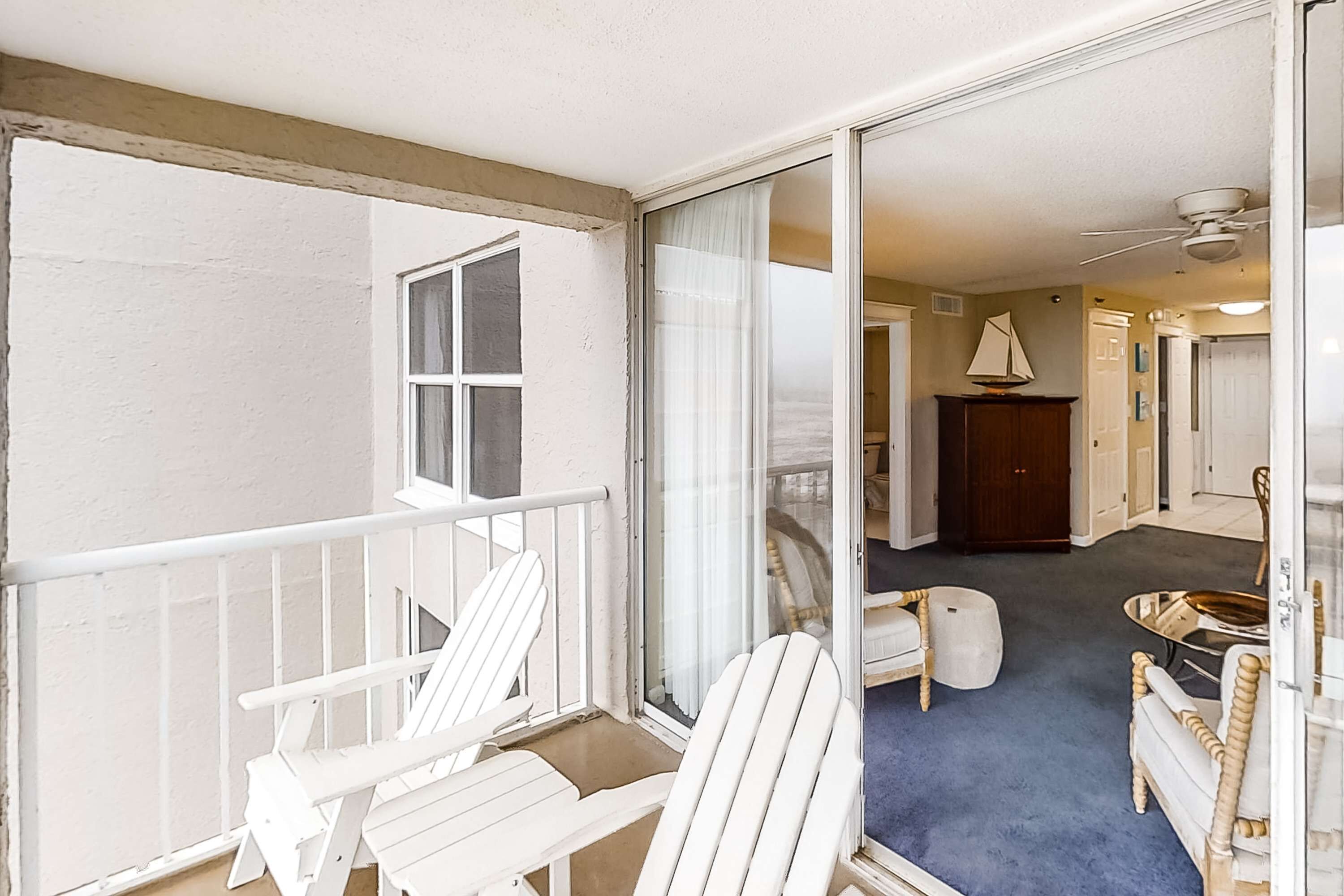 Un balcon avec des fauteuils Adirondack blancs, une porte-fenêtre coulissante donnant sur un salon doté d’une moquette bleue, d’une décoration sur le thème de la voile et d’un ventilateur de plafond.