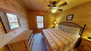 A wooden cabin bedroom with a bed, two windows, a ceiling fan, and a nightstand. - Harvest Moon Cottages - Peacepipe   00088 (Rockbridge)