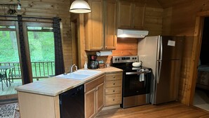 A wooden cabin kitchen with a dishwasher, stove, refrigerator, and a large window overlooking a forest. - Harvest Moon Cottages - Peacepipe   00088 (Rockbridge)
