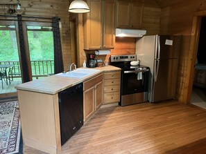 A wooden cabin kitchen with a dishwasher, stove, refrigerator, and a large window overlooking a forest. - Harvest Moon Cottages - Peacepipe   00088 (Rockbridge)