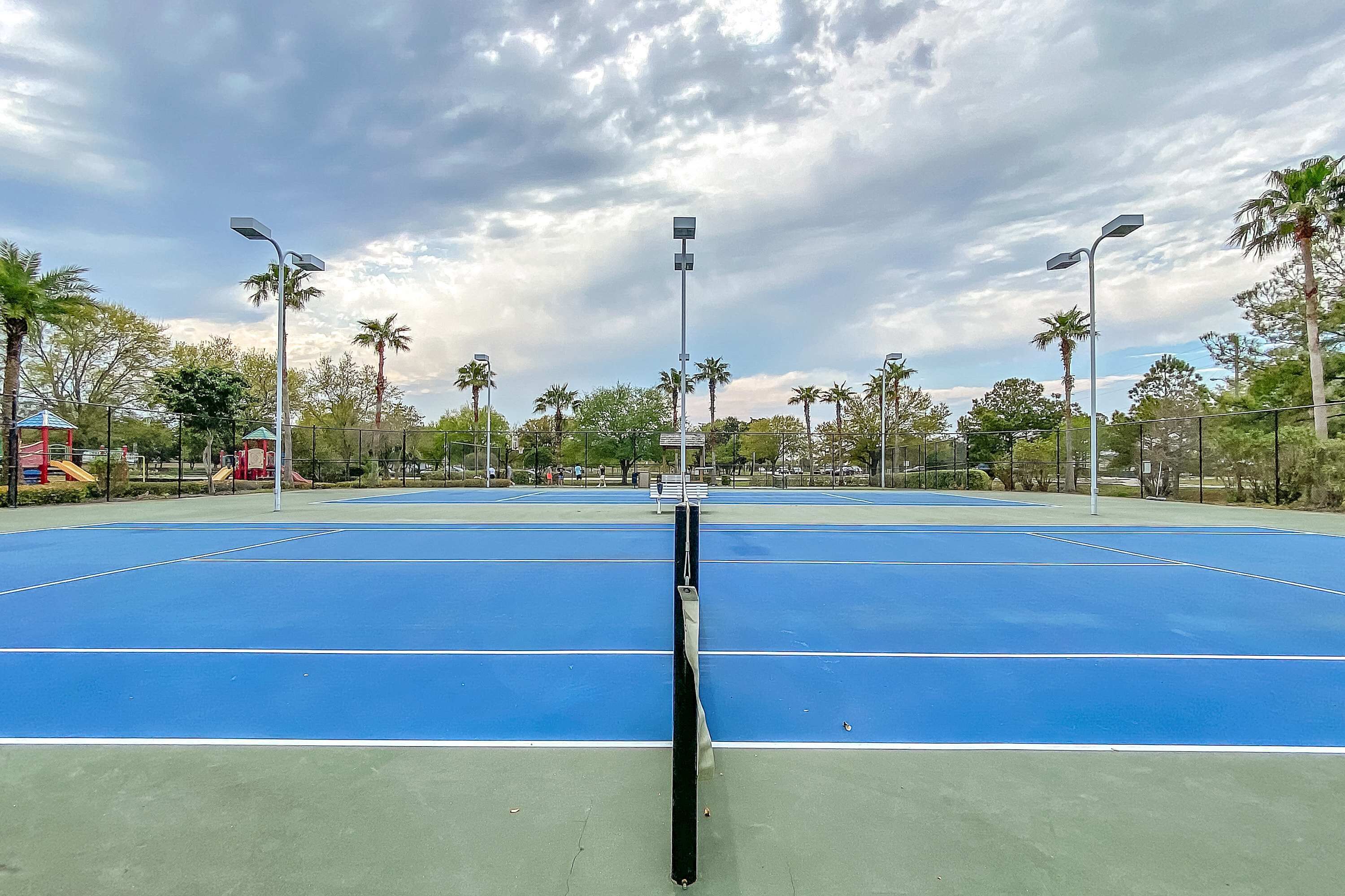 A tennis court with a net in the center, surrounded by palm trees and street lamps.