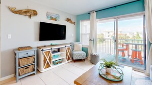 A living room with a TV, a small table with a plant, a chair, and a balcony with outdoor furniture. - Gulf view condo with pool, balcony, full kitchen, nautical theme, W/D, & AC (Destin)