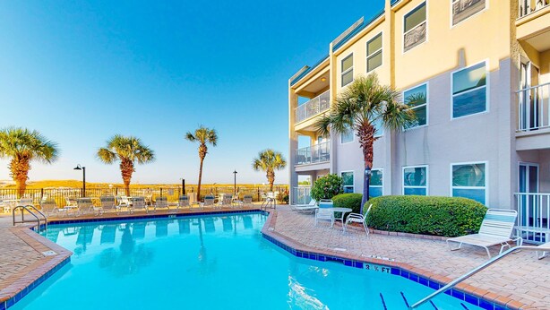A swimming pool with lounge chairs and palm trees, surrounded by a multi-story building with balconies. - Gulf view condo with pool, balcony, full kitchen, nautical theme, W/D, & AC (Destin)