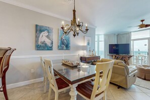 A dining area with a glass-top table, white chairs, a chandelier, and artwork on the walls. - Gorgeous & cozy beachfront condo with pool & beach access (Destin)