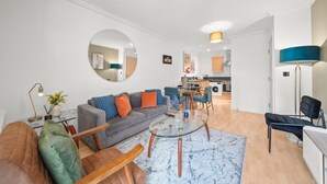 A modern living room with a grey sofa, a round coffee table, and a blue rug. There is a kitchen in the background with wooden cabinets and a dining area. - One bed apt opp tube station central line (London)