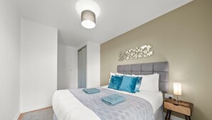 A neatly made bed with a grey patterned bedspread and two folded towels, a bedside lamp, and a wall-mounted abstract art piece. - One bed apt opp tube station central line (London)