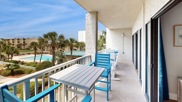 A balcony with blue chairs and a table, overlooking a pool and palm trees. - Snowbird-friendly condo with beach views, private beach, heated pool (Destin)