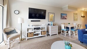 A living room with a sofa, a TV, a dining table with chairs, a clock, and a lamp. - Snowbird-friendly condo with beach views, private beach, heated pool (Destin)