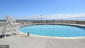 En rund pool med ett trädäck, en vit stol och utsikt över havet.