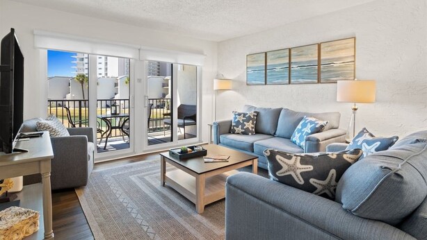 A modern living room with a sofa, coffee table, and a TV. There is a balcony with outdoor furniture and a view of the sky. - Ocean View 2BR Condo • Pets • Balcony • Sleeps 8 (Panama City Beach)