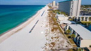 A beachfront hotel with a long sandy beach, a row of blue umbrellas, and a large building with multiple floors. - Ocean View 2BR Condo • Pets • Balcony • Sleeps 8 (Panama City Beach)