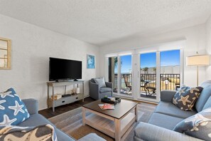 Un salon doté d’un téléviseur à écran plat, de canapés gris, d’une table basse et d’un balcon aménagé avec du mobilier d’extérieur.