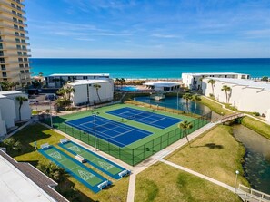 Un court de tennis entouré de bâtiments, avec une piscine à proximité et une vue sur l’océan.