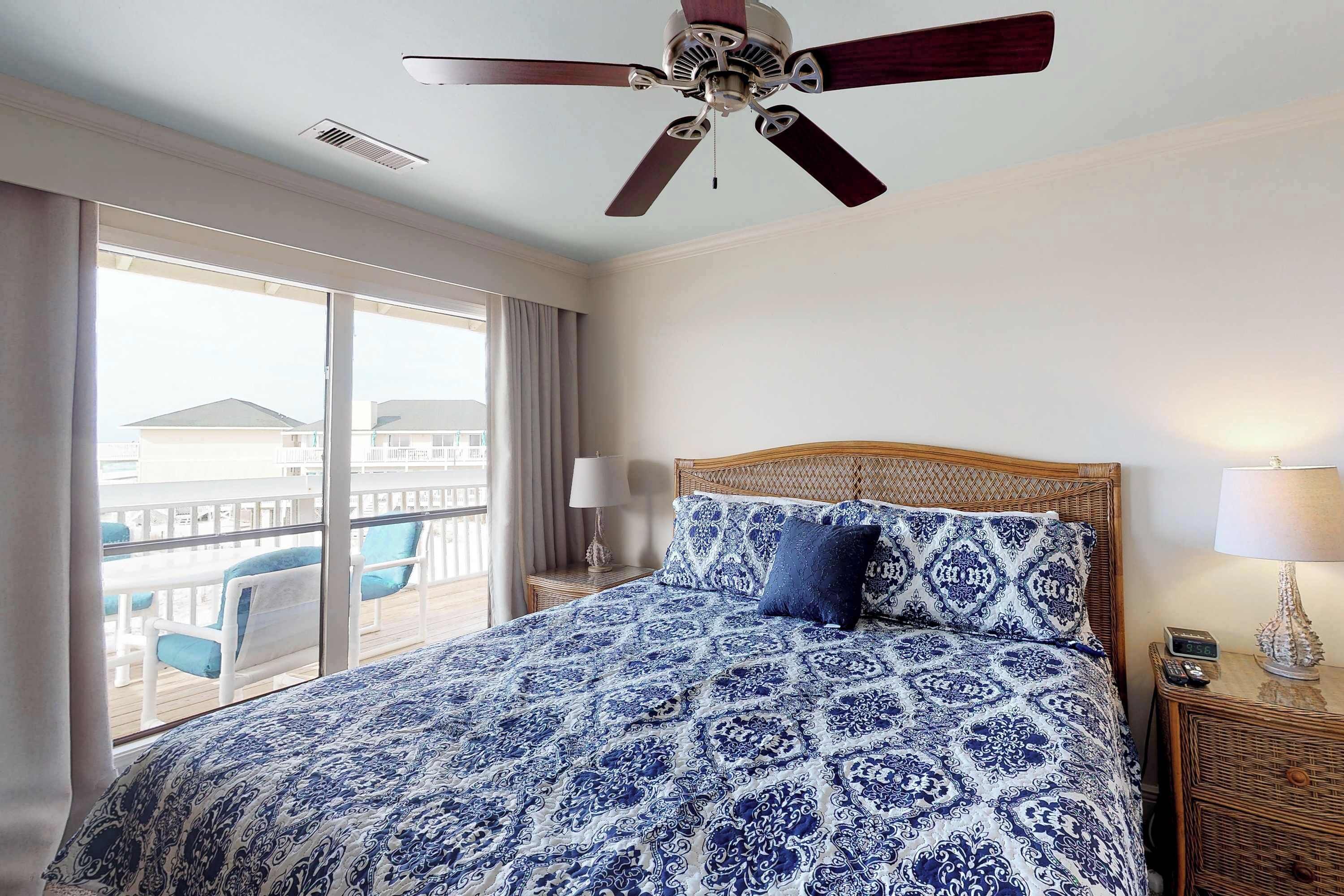 A hotel room with a large bed, a ceiling fan, a balcony with chairs, and a view of buildings.