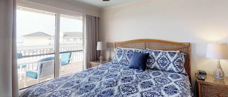 A hotel room with a large bed, a ceiling fan, a balcony with chairs, and a view of buildings.