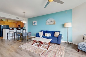 A modern living room with a blue wall, a blue sofa, and a dining area with wooden cabinets and a white table. - Resort condo with amazing pool, hot tub, tennis, fitness center & Wifi (Destin)
