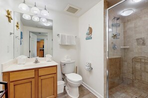 A bathroom with a wooden vanity, a toilet, a shower with a glass door, and a mirror with lights. - Resort condo with amazing pool, hot tub, tennis, fitness center & Wifi (Destin)