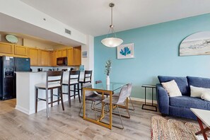 A modern kitchen with wooden cabinets, a black refrigerator, and a microwave. There is a dining area with a glass table and chairs, and a blue sofa in the living room. - Resort condo with amazing pool, hot tub, tennis, fitness center & Wifi (Destin)