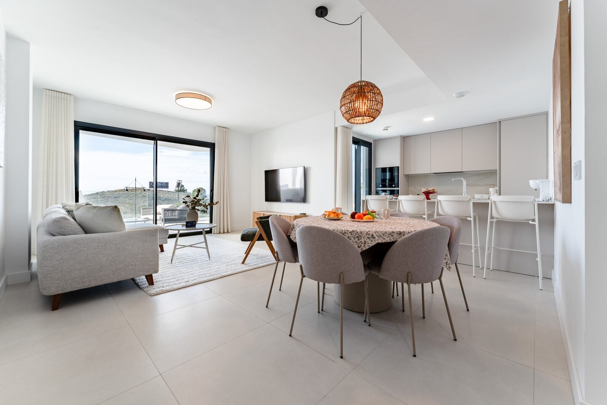 A modern living room with a sofa, dining table, and chairs, a kitchen area with white cabinets, and a large window offering a view.
