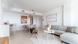 Ett modernt vardagsrum med en soffa, en matplats med stolar, ett köksområde och en hängande lampa.