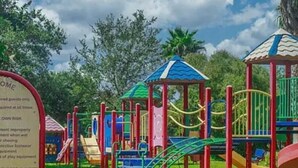 A playground with various play structures, including slides and climbing areas, surrounded by trees. - Snowbird-friendly condo with pool, hot tub, splash area, tennis, restaurant, gym (Destin)