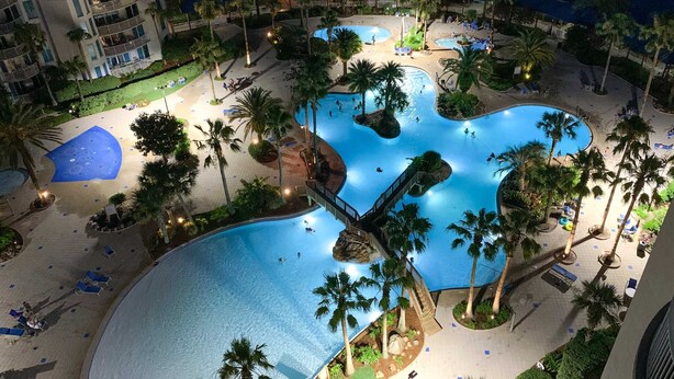 A large outdoor swimming pool surrounded by palm trees and lounge chairs, with a bridge connecting different sections of the pool. - Snowbird-friendly condo with pool, hot tub, splash area, tennis, restaurant, gym (Destin)