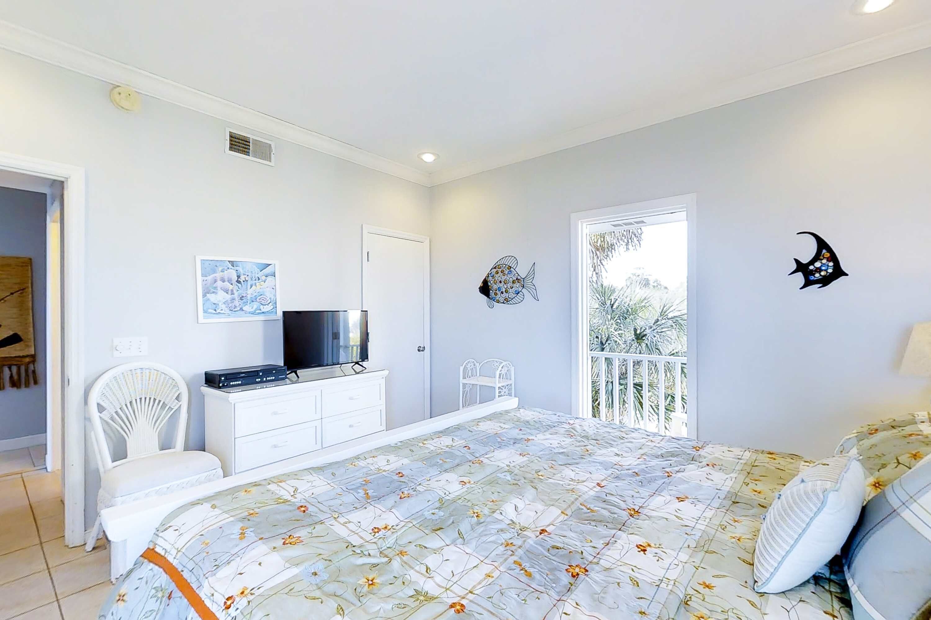 A bedroom with a bed, a TV, a white dresser, a chair, and a balcony with a view of trees.