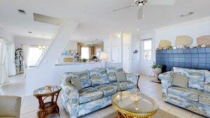 En rummelig stue med højt til loftet, en loftsvifte og en trappe, der fører til en etage ovenpå. Der er to sofaer, et glasbord og et lille sofabord.