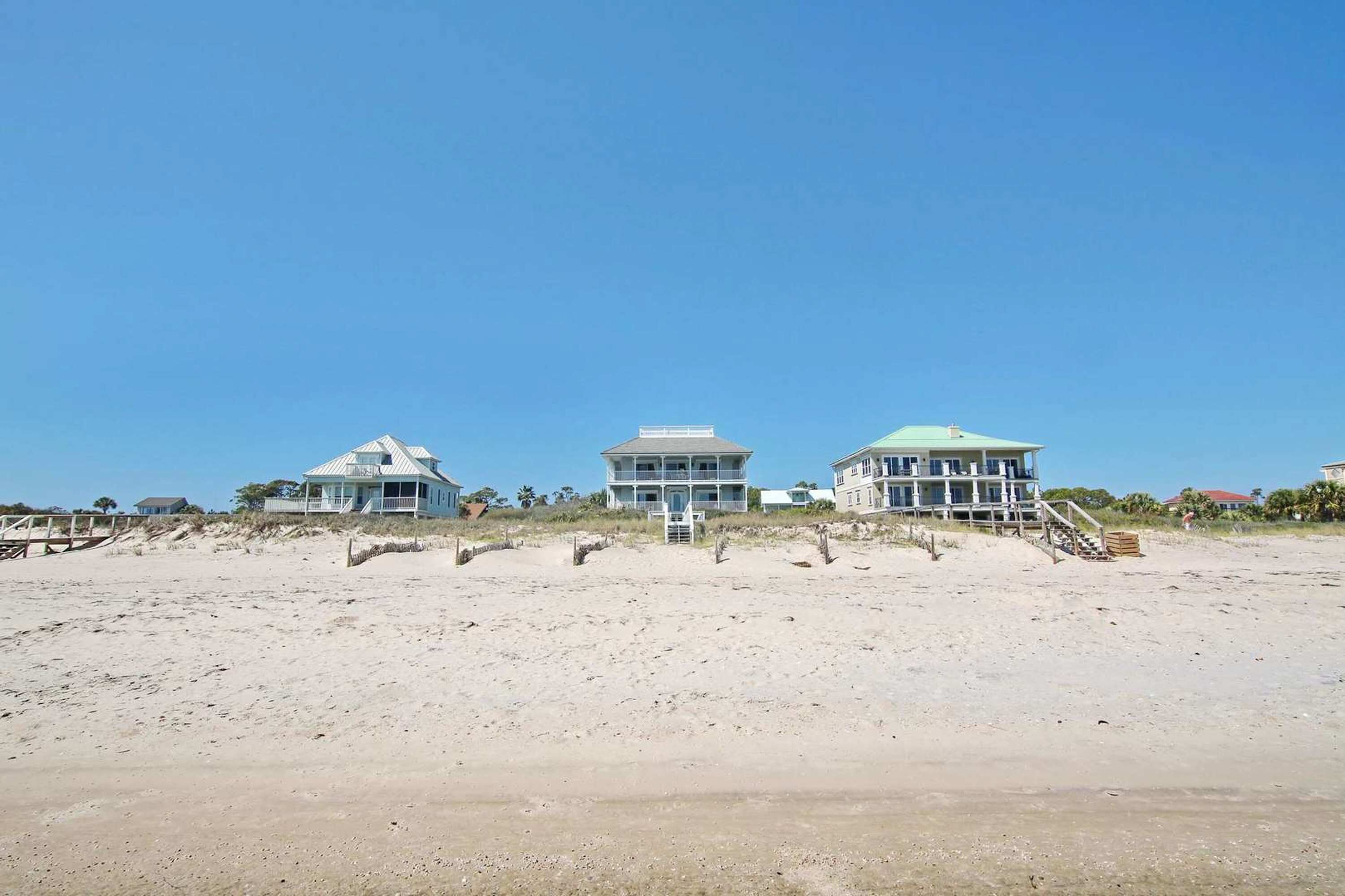 Een strandpand met drie grote huizen, elk in een eigen architectuur, gelegen aan een zandstrand onder een helderblauwe lucht.